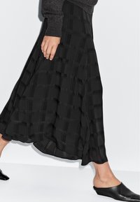 Vrouw die loopt in een zwarte geruite midi-rok met zwierige beweging, donkere trui en zwarte instap lage hakken tegen een witte achtergrond.