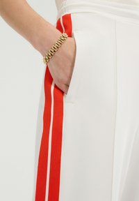 Pantalones blancos con una franja lateral en rojo y blanco. Presentan un bolsillo delantero y una tela suave y estructurada. Brazalete dorado en la muñeca.