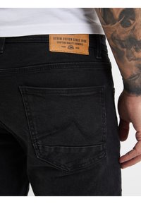Zwarte denim jeans met een patchlabel waarop "DENIM DRIVEN SINCE 1989" staat. Bevat gestikte zakken en een close-up van de textuur van de stof.