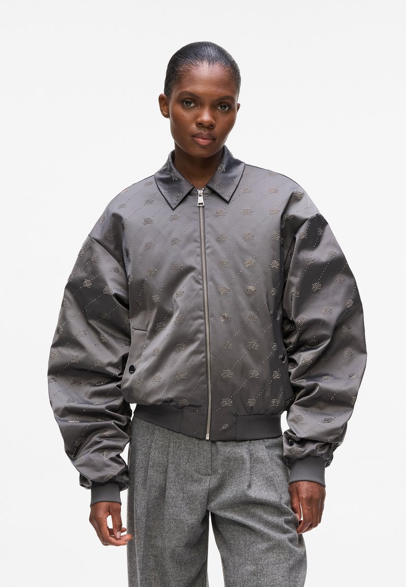 Giacca bomber grigia con maniche oversize, dettagli metallici e motivo logo ricamato. Presenta una chiusura con zip e un design del colletto.