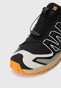 Black athletic shoe with textured fabric upper, orange rubber sole, and grey accents. Features a quick-lace system and reinforced toe cap.