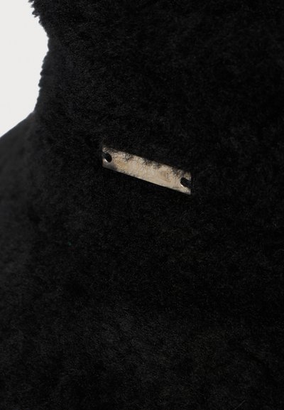 Close-up van zwart donzig stof met een klein rechthoekig metalen label dat aan de zijkanten door twee gaatjes is bevestigd.