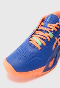 Blue athletic shoe with orange accents, mesh upper, textured surface, and green lace loops. Features a rubber sole and brand logo.