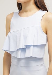 Světle modrý top bez rukávů s vrstvenými volány v oblasti prsou, vyrobený z hladké, mírně strukturované tkaniny; přiléhavý v pase.