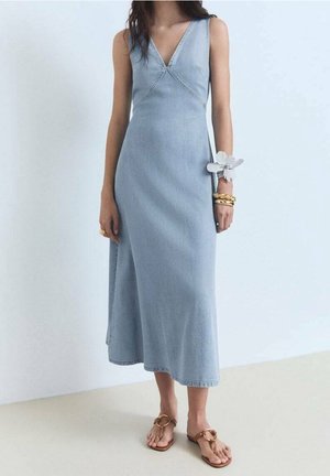 Femme portant une robe midi en denim bleu clair sans manches avec un décolleté en V, des bracelets dorés et des sandales marron avec des détails en chaîne.