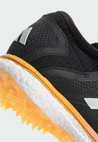 Černá sportovní bota s texturovaným síťovaným svrškem, gumovou oranžovou podrážkou, kontrastními bílými akcenty a detailním prošitím podél okrajů.