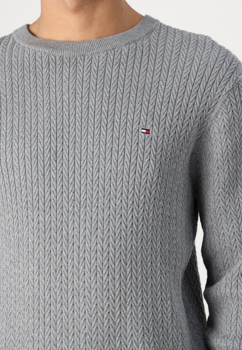 Pull en tricot gris avec un motif en chevron texturé, un col côtelé et un petit logo coloré sur la poitrine.