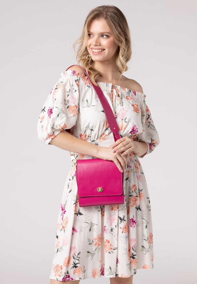 Borsa a spalla in pelle rosa con chiusura a ribalta, hardware dorato e tracolla regolabile. Esibita su un vestito a fantasia floreale.