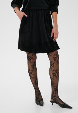 Femme portant une jupe en velours noire, des collants en dentelle florale noirs et des chaussures noires pointues à talons bas, debout, main dans la poche.