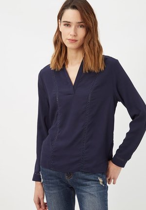 Mujer con blusa azul marino de manga larga con costuras decorativas y vaqueros azules desgastados, de pie con una mano en la cadera.