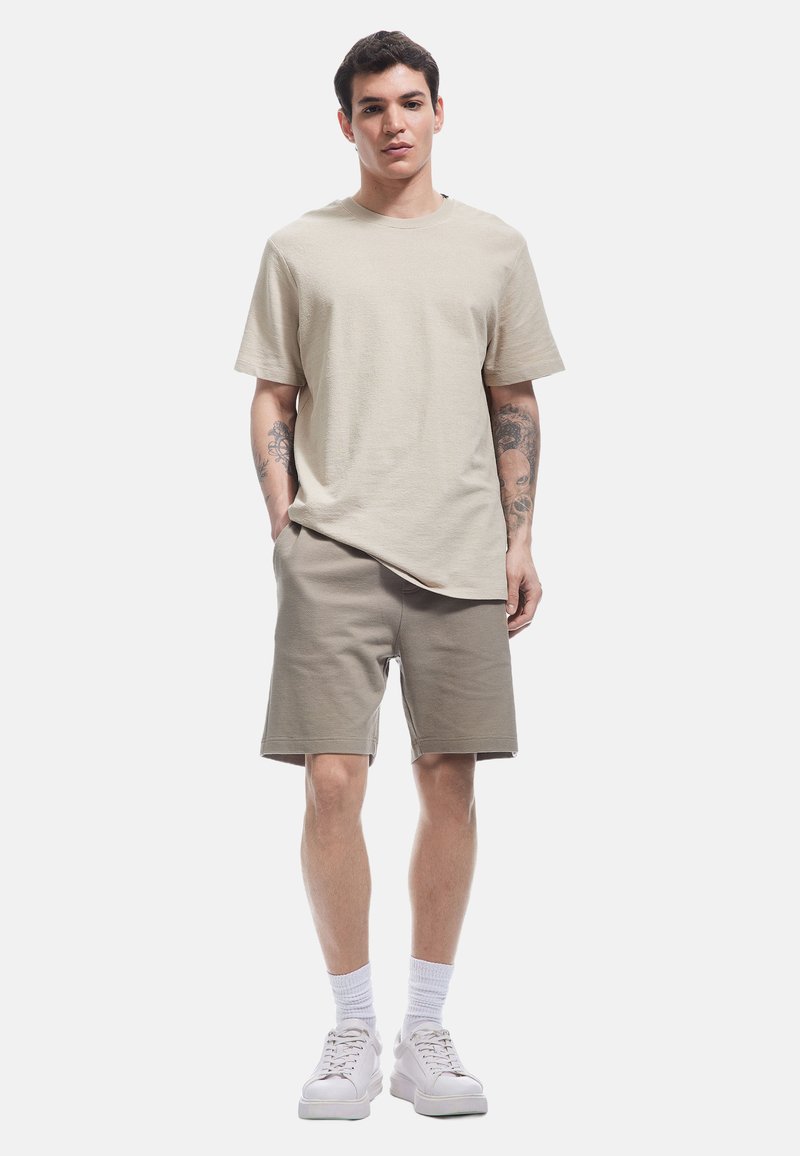 Camicetta beige a maniche corte abbinata a pantaloni corti grigio chiaro. Sneakers bianche e calzini corti. Design semplice, vestibilità rilassata, tessuto testurizzato.