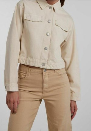 Kobieta ubrana w jasnobeżową krótka kurtkę dżinsową z srebrnymi guzikami oraz beżowe spodnie z wysokim stanem na szarym tle.