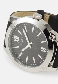 Guess Armbanduhr mit einem Gehäuse aus silbernem Edelstahl, schwarzem gemustertem Zifferblatt, weißen Stundenmarkierungen und einem schwarzen Lederarmband.