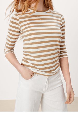 Person iført en beige og hvid stribet langærmet skjorte med den ene hånd i lommen på hvide bukser, stående mod en ensfarvet baggrund.