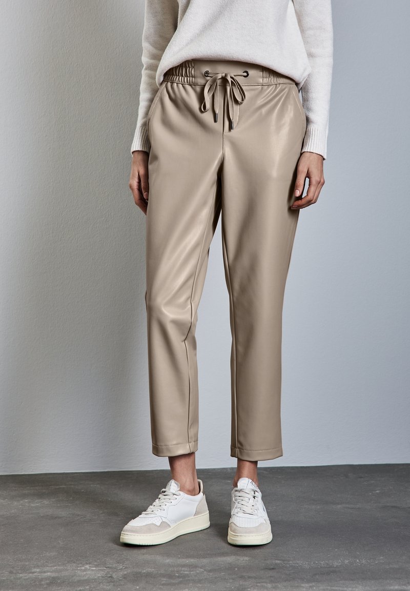 Femme portant un pantalon beige en simili-cuir avec cordon de serrage, des baskets blanches et un pull crème, debout sur un sol gris contre un mur uni.