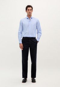 Lichtblauw button-up shirt met verticale strepen, gecombineerd met donkerblauwe broek en zwarte schoenen. Effen achtergrond, staande pose.