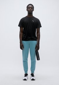 Μαύρο αθλητικό μπλουζάκι με κοντά μανίκια, ανοιχτό μπλε παντελόνι φόρμας, μαύρα παπούτσια, κρατώντας ένα μαύρο μπουκάλι νερού, στέκεται μπροστά σε απλό φόντο.