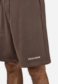 Brune bomullsshorts med elastisk midjebånd, sidelommer og en hvit "PEGADOR"-logo på høyre bein. Glatt tekstur, kne-lengde design.