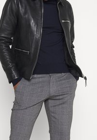 Schwarze Lederjacke mit Reißverschluss, kombiniert mit einem navyblauen Pullover und grauen karrierten Hosen, die ein strukturiertes Muster und Seitentaschen aufweisen.