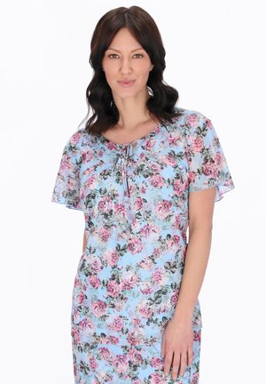 Žena s tmavými vlasy po ramena, oblečená v světle modrých šatech s růžovým květinovým vzorem, stojící před bílým pozadím.