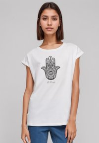 Weißes Baumwoll-T-Shirt mit kurzen, gerollten Ärmeln; zeigt ein schwarzes Handgrafikdesign mit komplizierten Mustern und einem Augensymbol.