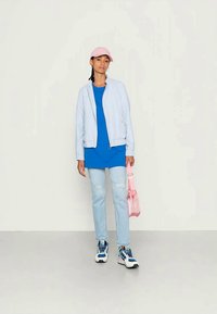 Young person wearing a pink cap, light blue jacket, blue shirt, ripped light jeans, sneakers, and holding a pink bag against a plain white background.