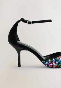 Scarpa alta in velluto nero con cinturino alla caviglia e decorazioni in paillettes multicolori sulla punta. Design elegante e affusolato con tacco lucido.