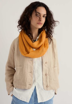 Mujer con cabello rizado y oscuro que lleva una chaqueta beige, camisa blanca, pantalones azules y una bufanda amarilla mostaza sobre un fondo liso.