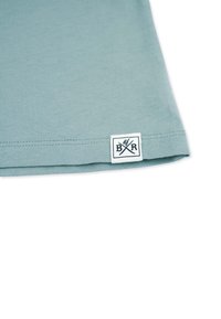 Lys blå bomulds T-shirt med en syet kant og et rektangulært mærke med "B R" og krydsede redskaber i marineblå.