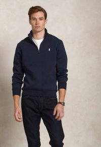 Jūras spēku zipsweaters, izgatavots no mīkstas auduma, ar stāvošu apkakli, ribotām aprocēm un mazu izšūtu logo uz krūtīm. Apvienots ar tumšiem džinsiem.