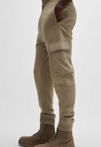 Khaki Joggerhosen mit elastischem Bund, seitlichen Taschen, Cargo-Tasche und gerippten Bündchen. Kombiniert mit braunen Schnürstiefeln.