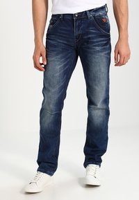 Donkerblauwe denim jeans met een vervaagde textuur, rechte pijpen en zichtbare stiksels. Gecombineerd met witte sneakers.