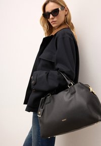 Svart läderhandväska med gyllene detaljer. Har en texturerad yta, dubbla handtag och en dekorativ etikett. Bärs med en mörk jacka och solglasögon.