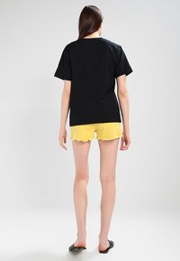 Camiseta de algodón negra combinada con shorts amarillos deshilachados, con un corte relajado y mangas cortas. El modelo lleva sandalias negras.