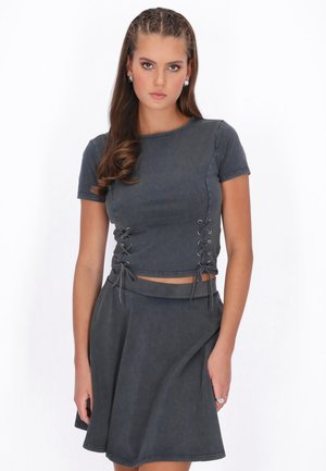 Tânără cu părul lung, împletit și ondulat, purtând un top scurt cu mâneci scurte, de culoare gri închis, cu șireturi laterale, și o fustă evazată asortată.