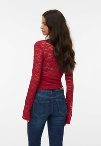 Rode kant blouse met lange mouwen, een getailleerde snit en uitlopende mouwen, gecombineerd met donkerblauwe jeans. De textuur is delicaat en doorschijnend.