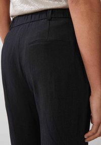 Mustat räätälöidyt housut, joissa on teksturoitu kangas, takatasku ja rento istuvuus. Materiaali vaikuttaa kevyeltä ja sileältä.