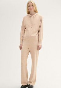 Felpa con cappuccio beige lavorata a maglia con cordini, abbinata a pantaloni larghi coordinati. Texture morbida, vestibilità casual, design minimalista. Calzature nere.