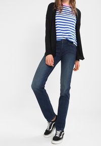 Kobieta ubrana w czarny kardigan, niebiesko-biały w paski koszulę, niebieskie dżinsy i czarne sneakersy, stoi z jedną nogą skrzyżowaną.