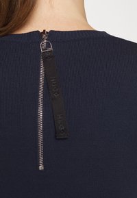Haut côtelé navy avec fermeture éclair au dos, dotée d'une tirette en tissu noir étiquetée "HUGO". Texture lisse et ajustée sur l'ensemble.
