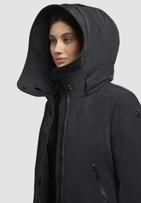 Frau in schwarzer Winterjacke mit großer Kapuze, durchgehendem Reißverschluss und seitlicher Reißverschlusstasche, die leicht nach rechts schaut.