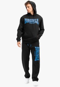 Zwart hoodie en trainingsbroek set, met blauwe "Lonsdale London" branding. Hoodie heeft een voorzak; de broek heeft een elastische tailleband en een trekkoord.