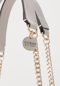 Grå lædertaske-strop med gyldne kæde-detaljer og et rundt metalmærke med "GUESS EST. 1981" præget. Synlige syninger.