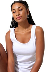 White sleeveless top made of cotton, features a scoop neckline and relaxed fit. The model has braided hair and gold hoop earrings.