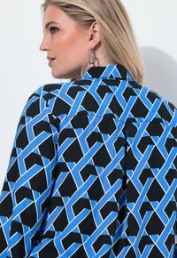 Sort bluse med et blåt og hvidt geometrisk mønster, med en klassisk krave og knapdetaljer bagpå. Glat stofstruktur.