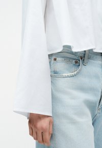 Χέρι που ακουμπάει στο πλάι, φορώντας λευκό μακρυμάνικο μπλουζάκι με φαρδιές μανσέτες και ανοιχτόχρωμα τζιν παντελόνια με εμφανείς λεπτομέρειες μπροστινών τσεπών.