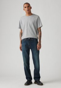 Grijze T-shirt met korte mouwen, blauwe denim jeans met een lichte vervaging en zwarte schoenen. Zichtbare tatoeages op de armen. Effen achtergrond.