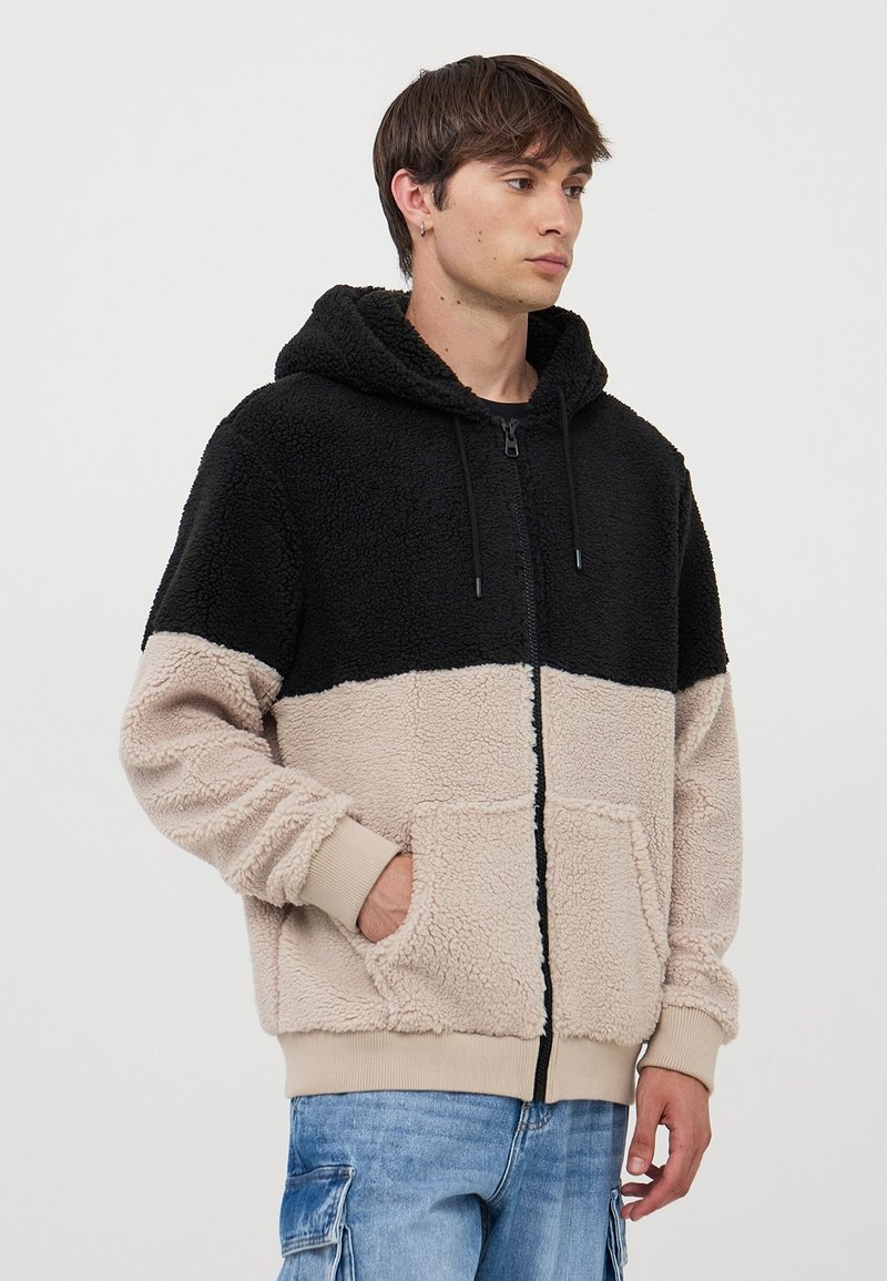 Giacca con cappuccio e zip, in morbido tessuto di peluche nero e beige, dotata di due tasche frontali e polsini a costine. Texture soffice con blocco di colore a contrasto.
