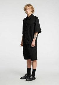 Camisa negra de manga corta con botones y bolsillo en el pecho, combinada con pantalones cortos negros holgados y mocasines negros, llevados con calcetines negros.