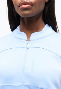 Κοντινό πλάνο ενός ατόμου που φοράει ανοιχτό γαλάζιο τοπ με φερμουάρ και μικρό λογότυπο Calvin Klein στο στήθος, εμφανίζοντας τους ώμους και τα χείλη.